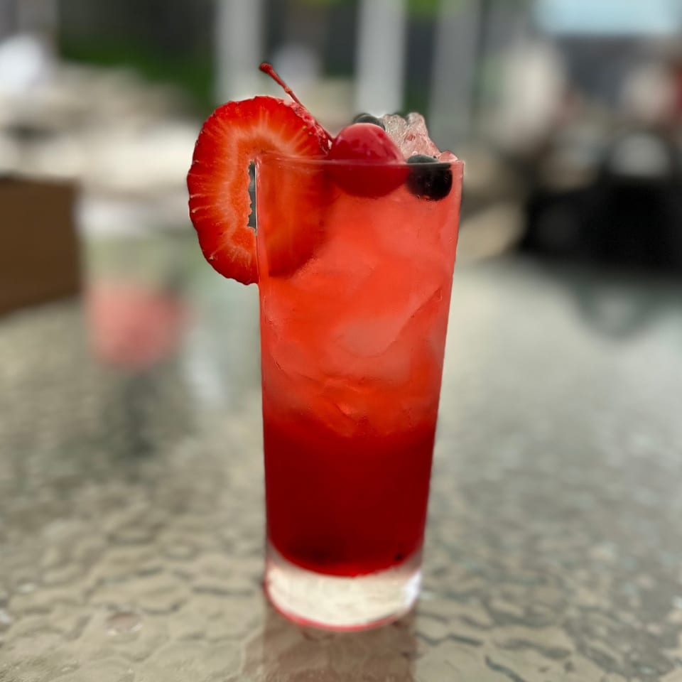A vibrant red cocktail, garnished with a strawberry slice, cherry, and blueberries, on a textured table.