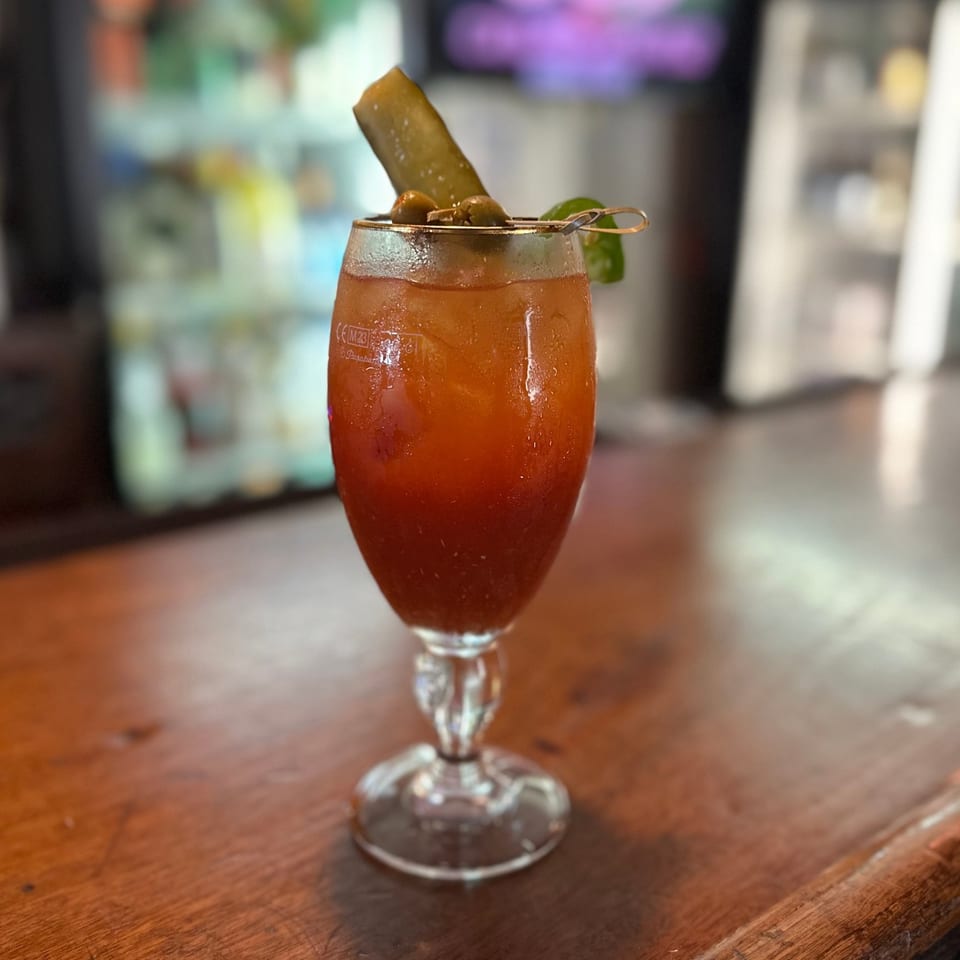 A Bloody Mary cocktail with a pickle spear and olives on a wooden bar top.