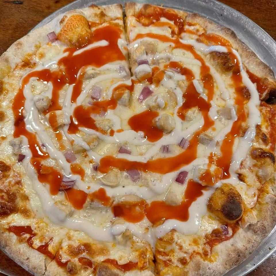 Buffalo chicken pizza with red sauce, white drizzle, chicken, and red onions on a metal tray.