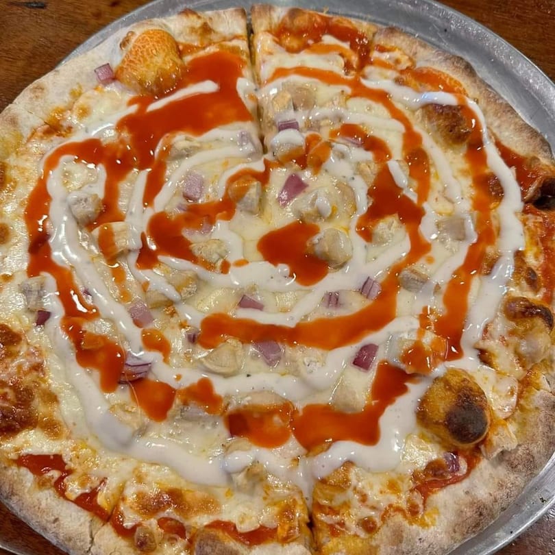 Buffalo chicken pizza with red sauce, white drizzle, chicken, and red onions on a metal tray.