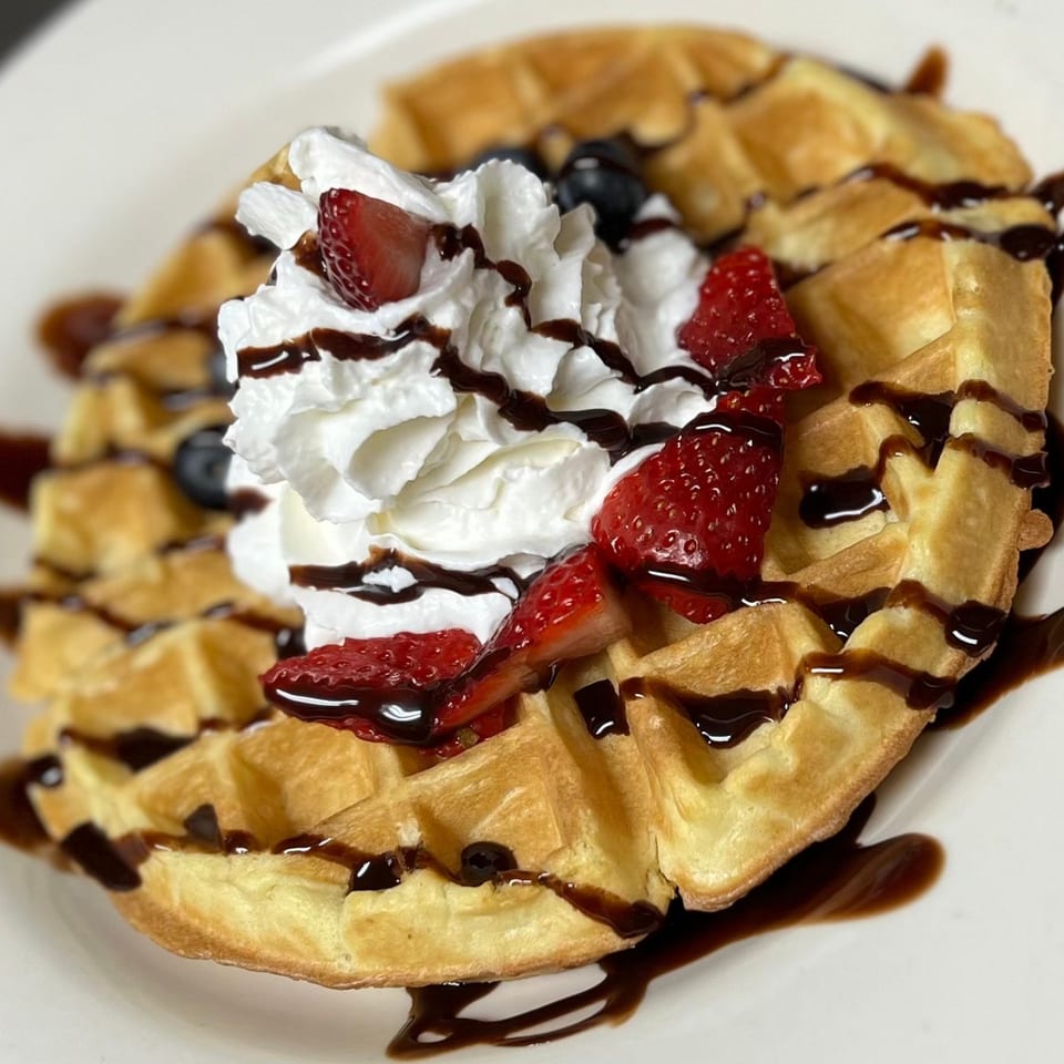 Golden waffle topped with whipped cream, fresh strawberries, blueberries, and chocolate drizzle on a white plate.
