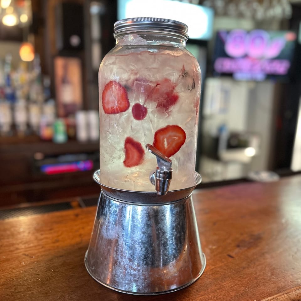A large glass drink dispenser with ice and sliced strawberries sits on a wooden bar.