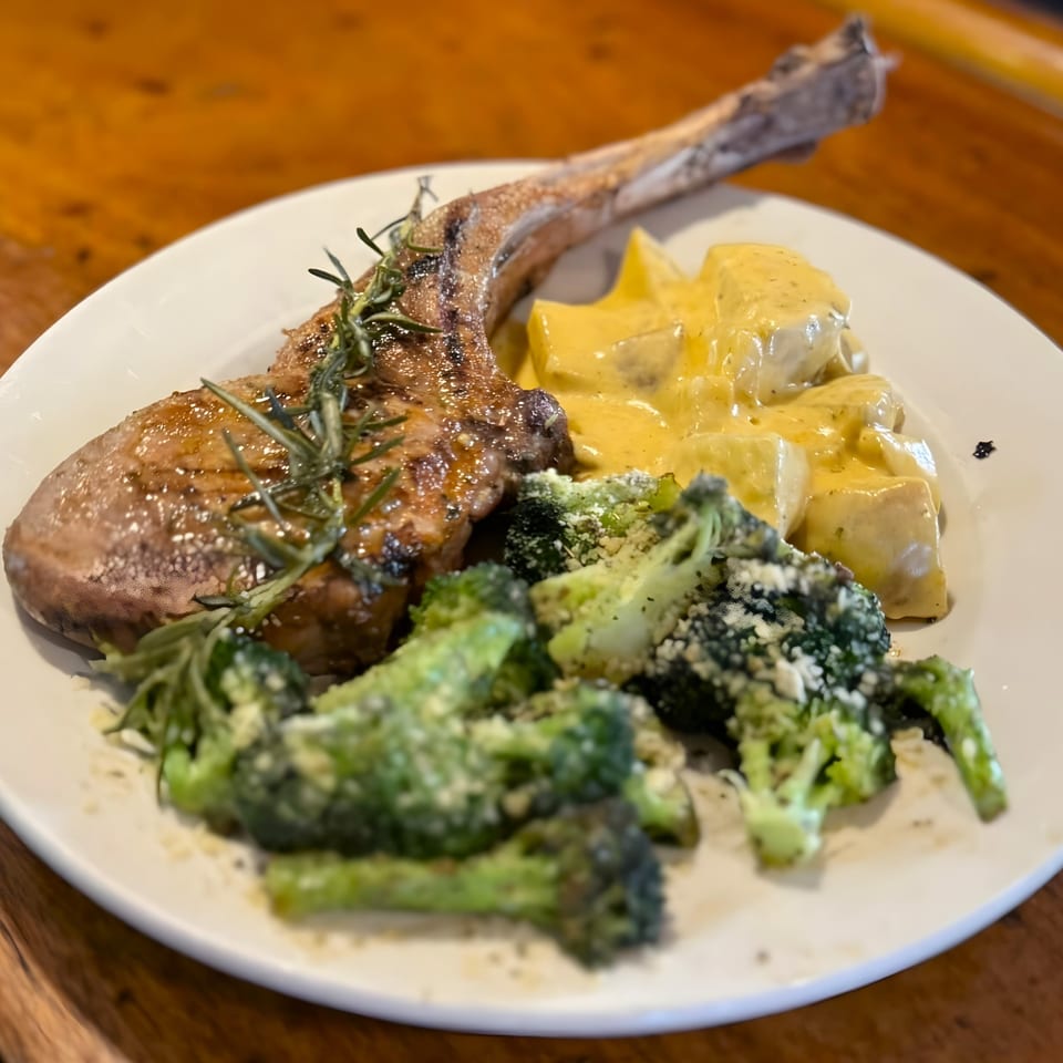 A bone-in pork chop with rosemary, cheesy potatoes, and parmesan-dusted broccoli on a white plate.