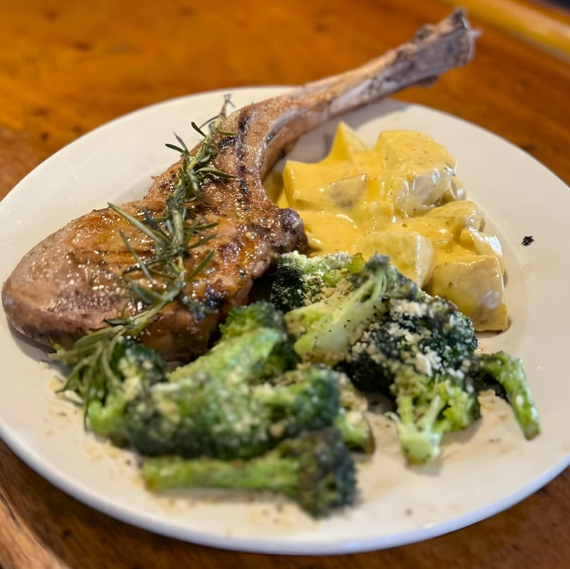 A bone-in pork chop with rosemary, cheesy potatoes, and parmesan-dusted broccoli on a white plate.