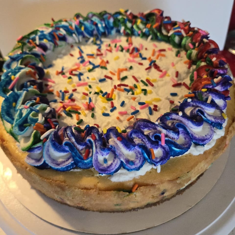 A round cake with white frosting, colorful sprinkles, and blue, purple, green, and red piped icing.
