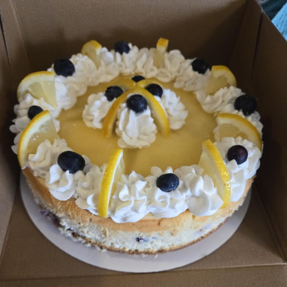 Lemon cheesecake with whipped cream, blueberries, and lemon slices in a brown box.