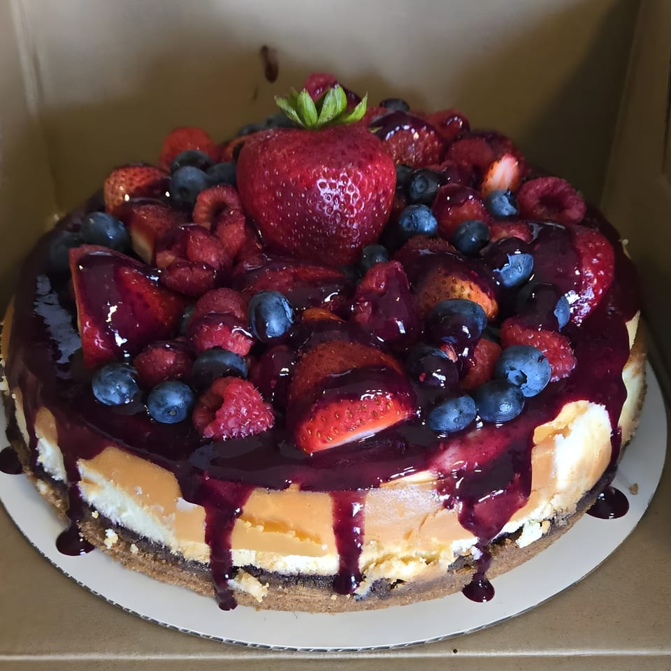 Cheesecake topped with strawberries, blueberries, raspberries, and a berry glaze in a cardboard box.
